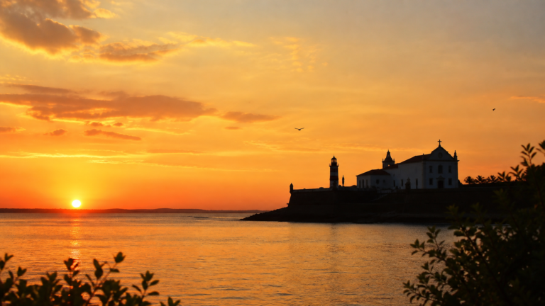 Sunset view over the Bay of All Saints from a point of land, with the silhouette of a colonial church and fort in Salvador, Bahia
