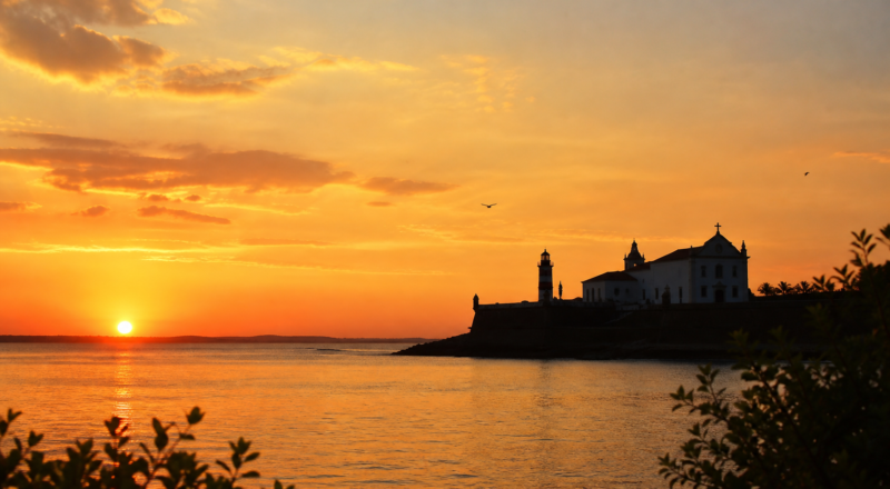 Sunset view over the Bay of All Saints from a point of land, with the silhouette of a colonial church and fort in Salvador, Bahia
