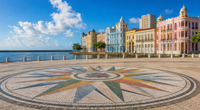 Sunny wide-angle view of Marco Zero in Recife Antigo featuring the colorful Rosa dos Ventos ground mosaic and historic waterfront buildings.