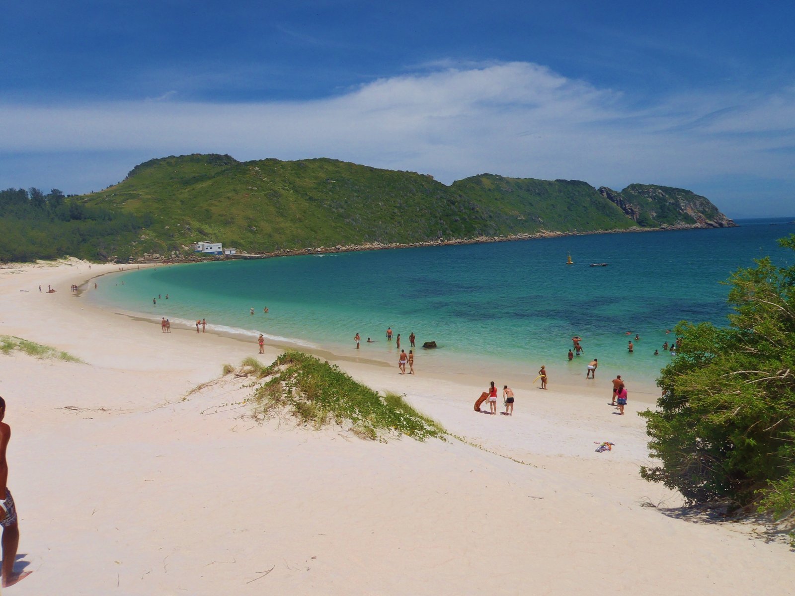 Praia do Farol, na Ilha do Cabo Frio, em Arraial do Cabo, com areia muito branca e dunas com vegetação rasteira em primeiro plano, mar verde-água transparente e banhistas na beira, e morros verdes da ilha ao fundo