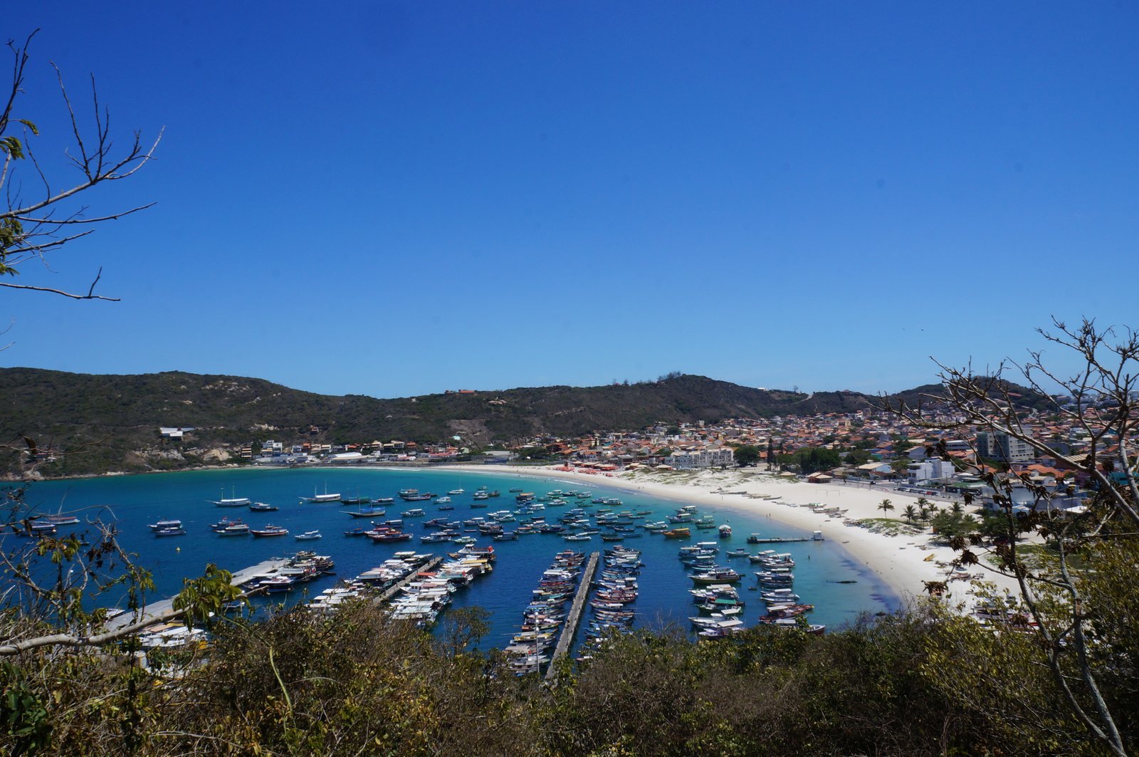 Vista do alto da Praia dos Anjos, em Arraial do Cabo, com dezenas de barcos de passeio coloridos atracados nos píeres da Marina dos Pescadores, a areia branca em curva e as casas da cidade atrás