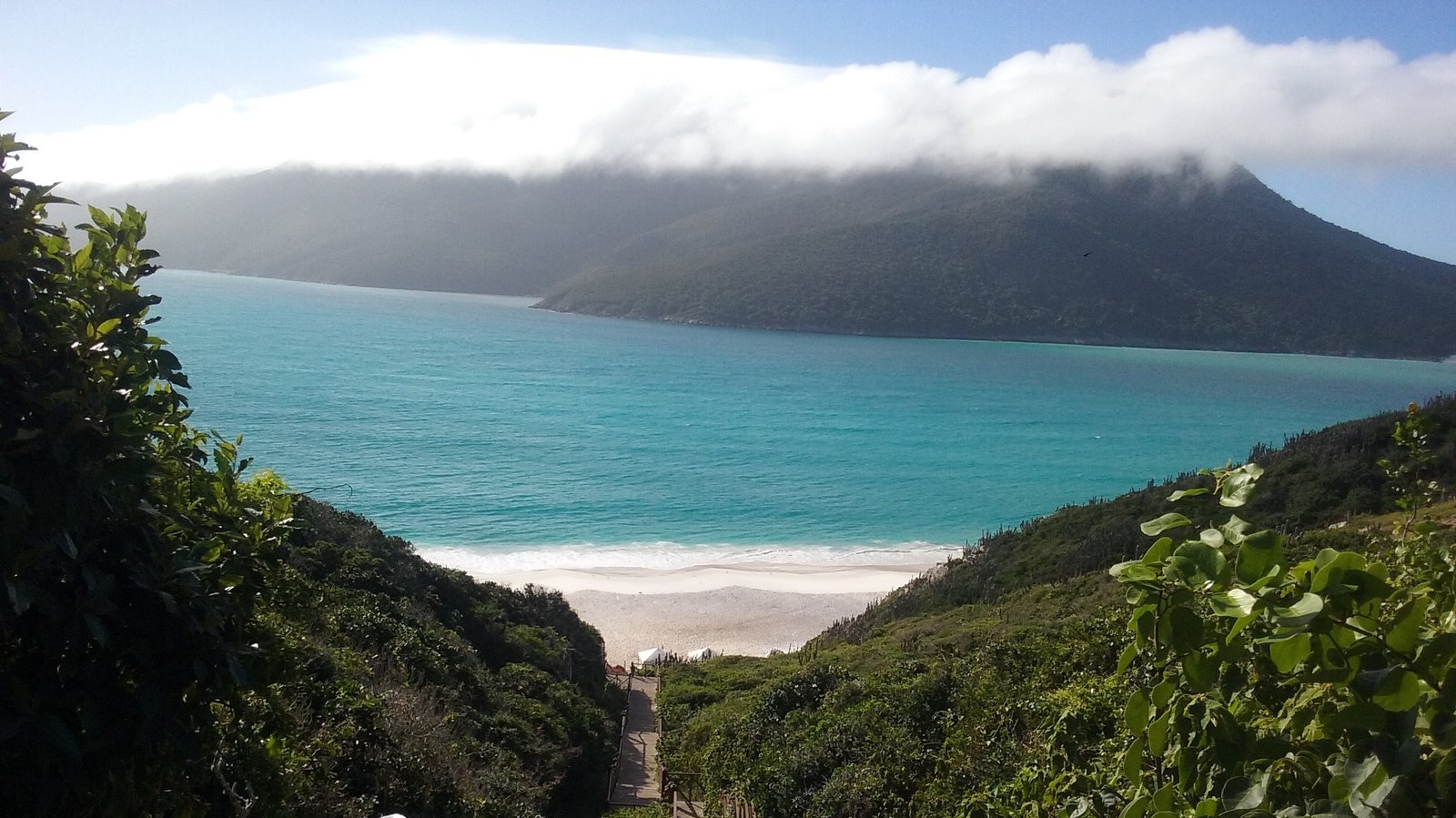 Escadaria descendo entre a vegetação do morro em direção às Prainhas do Pontal do Atalaia, em Arraial do Cabo, com a areia branca e o mar azul-esverdeado lá embaixo e a Ilha do Cabo Frio coberta de nuvens ao fundo