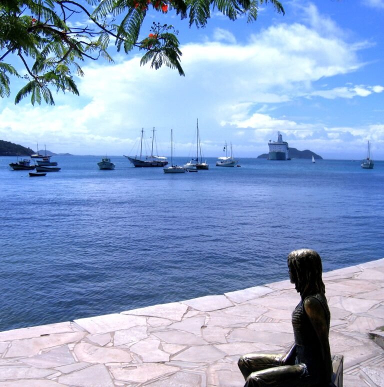 Estátua de bronze de Brigitte Bardot sentada numa mala, de frente para a baía da Armação, com barcos de pesca ancorados e morros verdes ao fundo