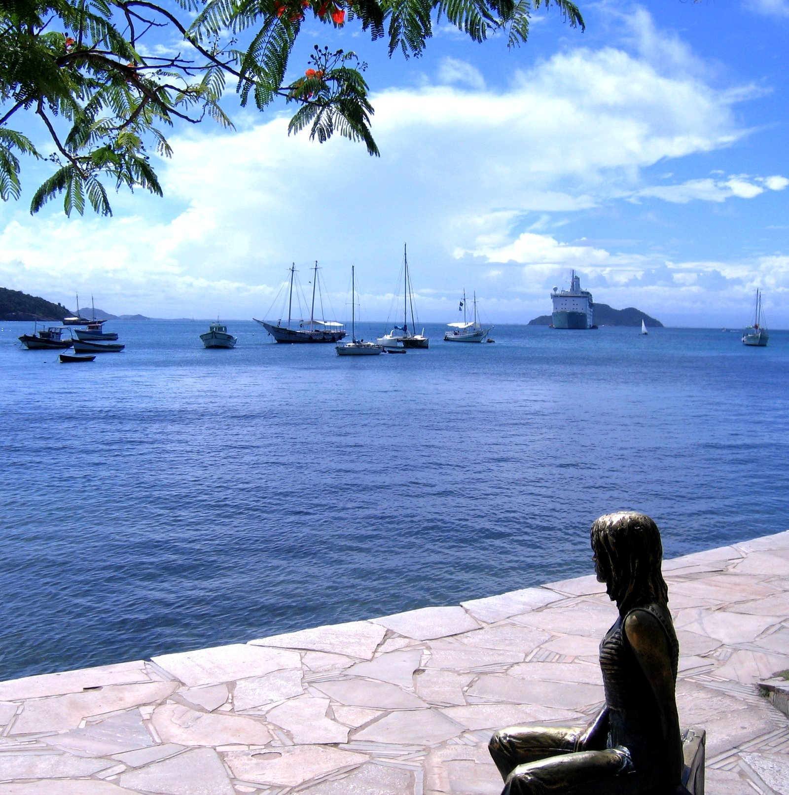 Estátua de bronze de Brigitte Bardot sentada numa mala, de frente para a baía da Armação, com barcos de pesca ancorados e morros verdes ao fundo