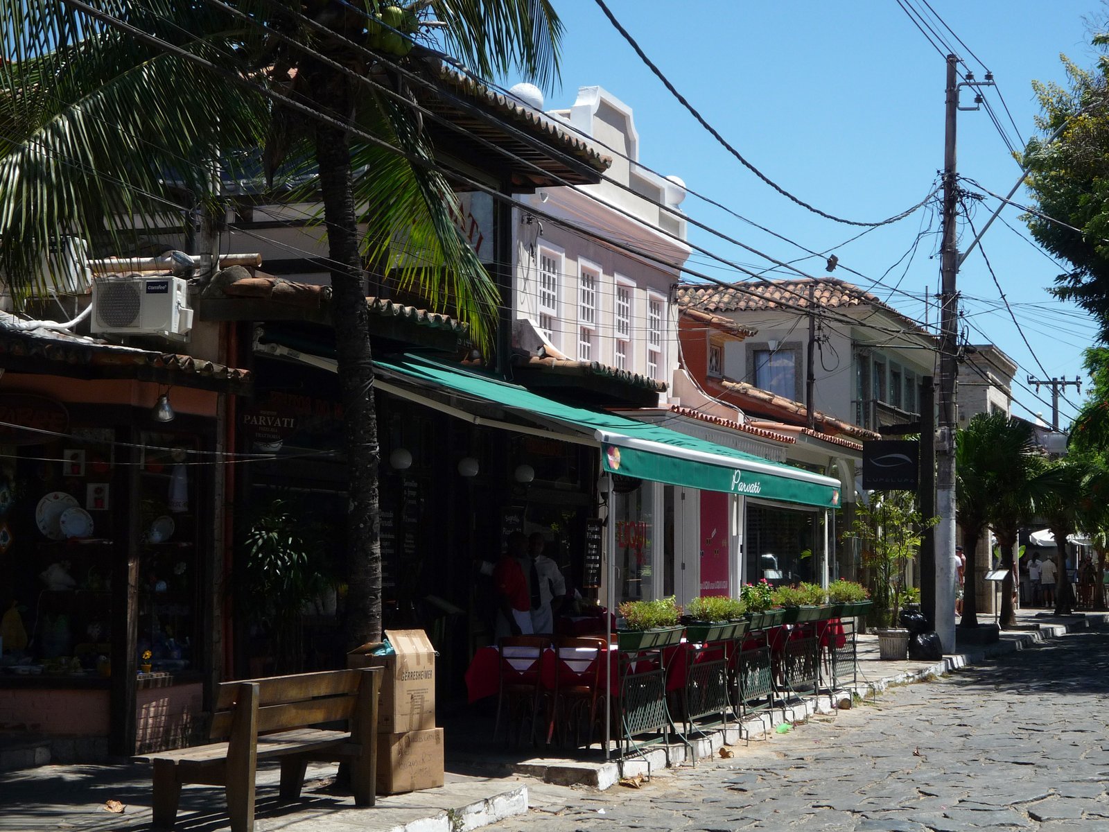 Rua das Pedras, em Búzios, com calçamento de pedras irregulares, casas baixas pintadas de branco e cores claras, lojas e pessoas caminhando sob céu azul