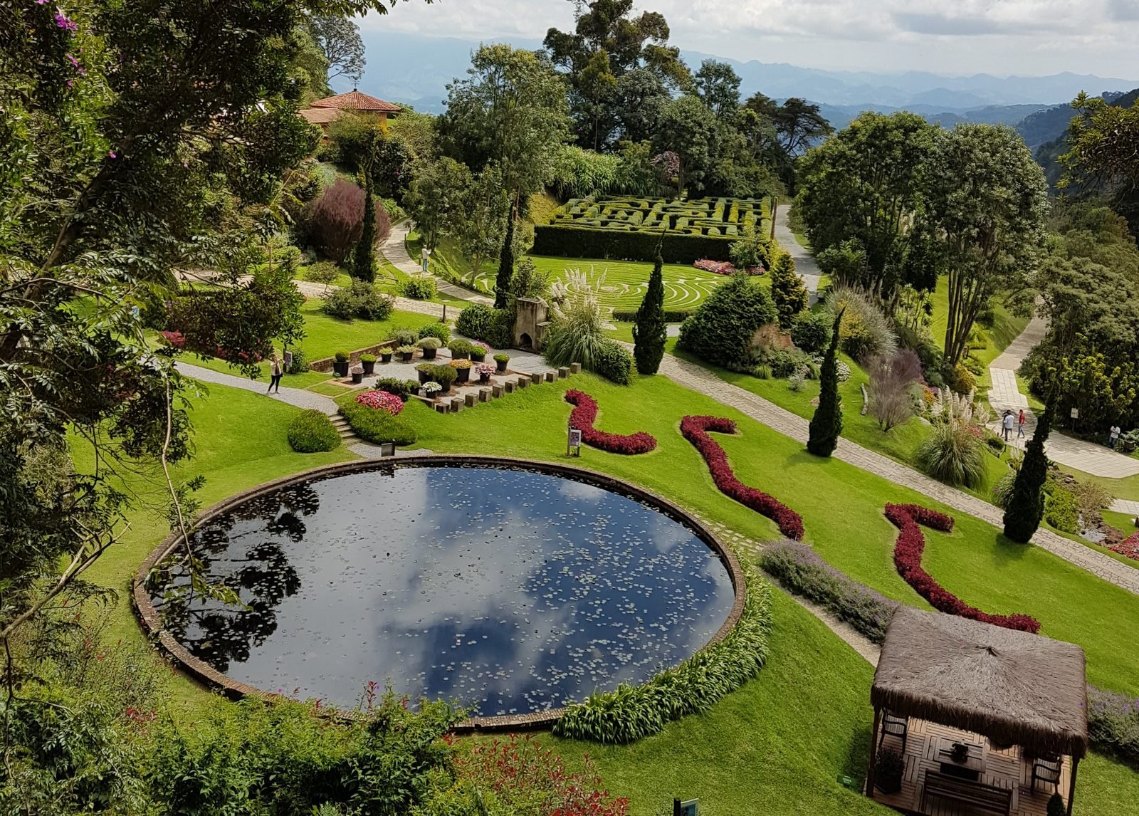 Vista do alto do Parque Amantikir, com um lago redondo espelhado, canteiros floridos em curvas, gramados e as montanhas da Serra da Mantiqueira ao fundo