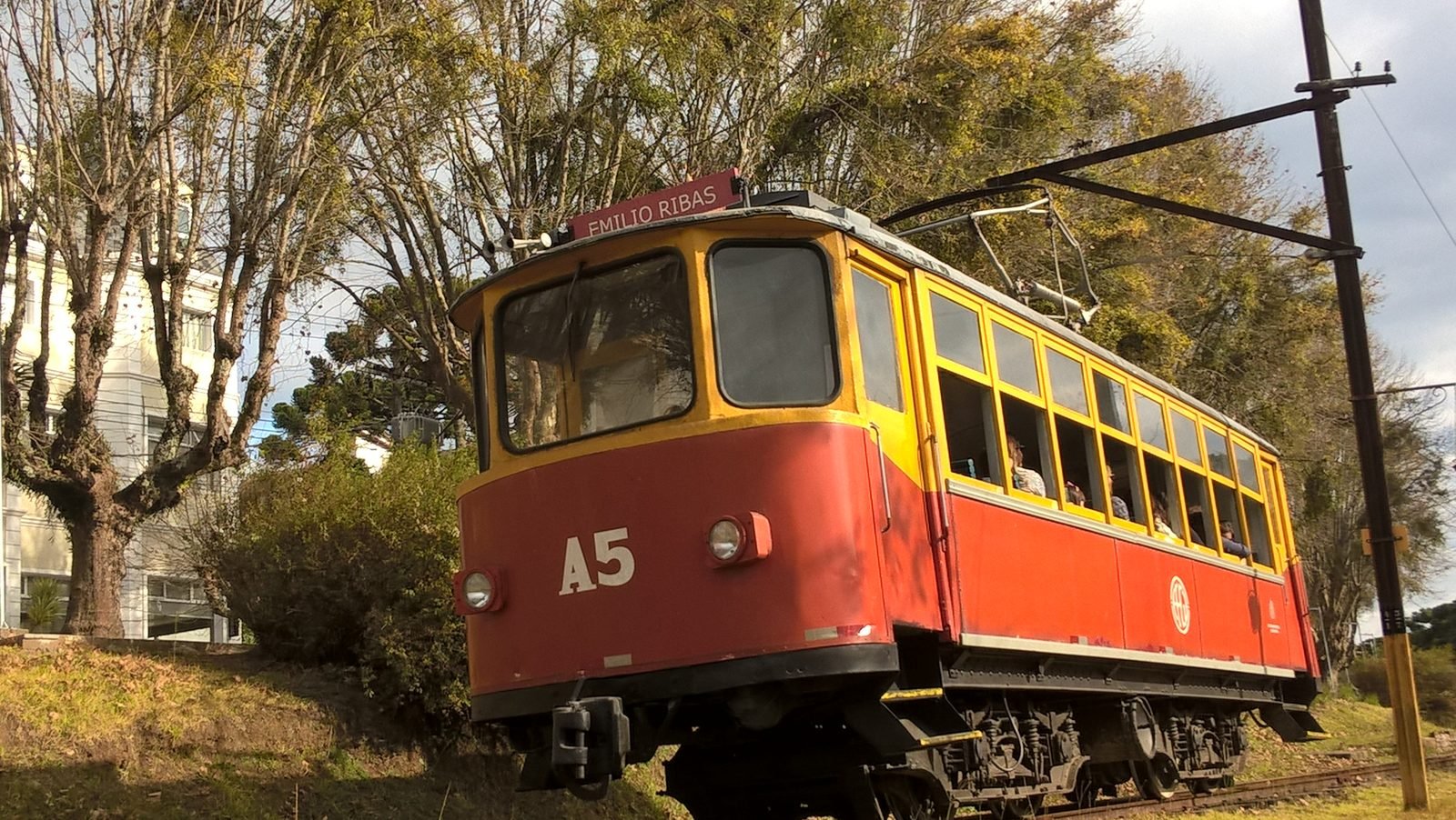 Bonde turístico vermelho e amarelo A5 da Estrada de Ferro Campos do Jordão, com a placa Emílio Ribas, nos trilhos entre árvores