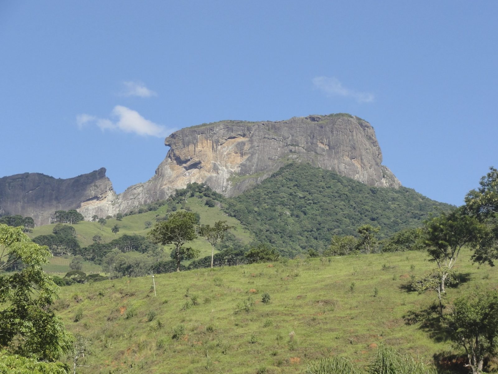As formações rochosas do Bauzinho e da Pedra do Baú, em São Bento do Sapucaí, acima de morros cobertos de mata e pasto verde sob céu azul