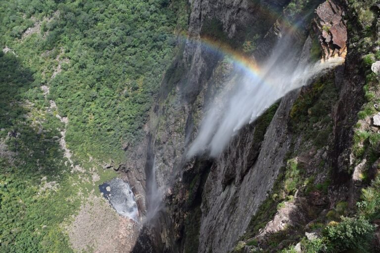Vista do alto da Cachoeira da Fumaça, com a água caindo por um paredão de pedra até o vale verde lá embaixo e um arco-íris formado na névoa