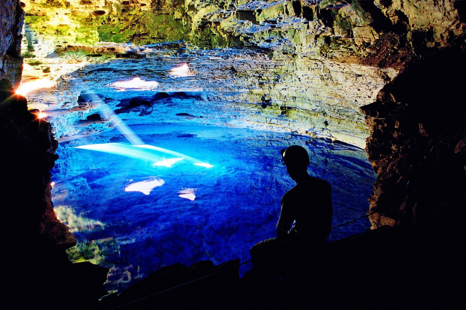 Feixe de luz do sol atravessando a caverna do Poço Encantado e tocando a água azul-turquesa, com a silhueta de uma pessoa sentada nas pedras à esquerda