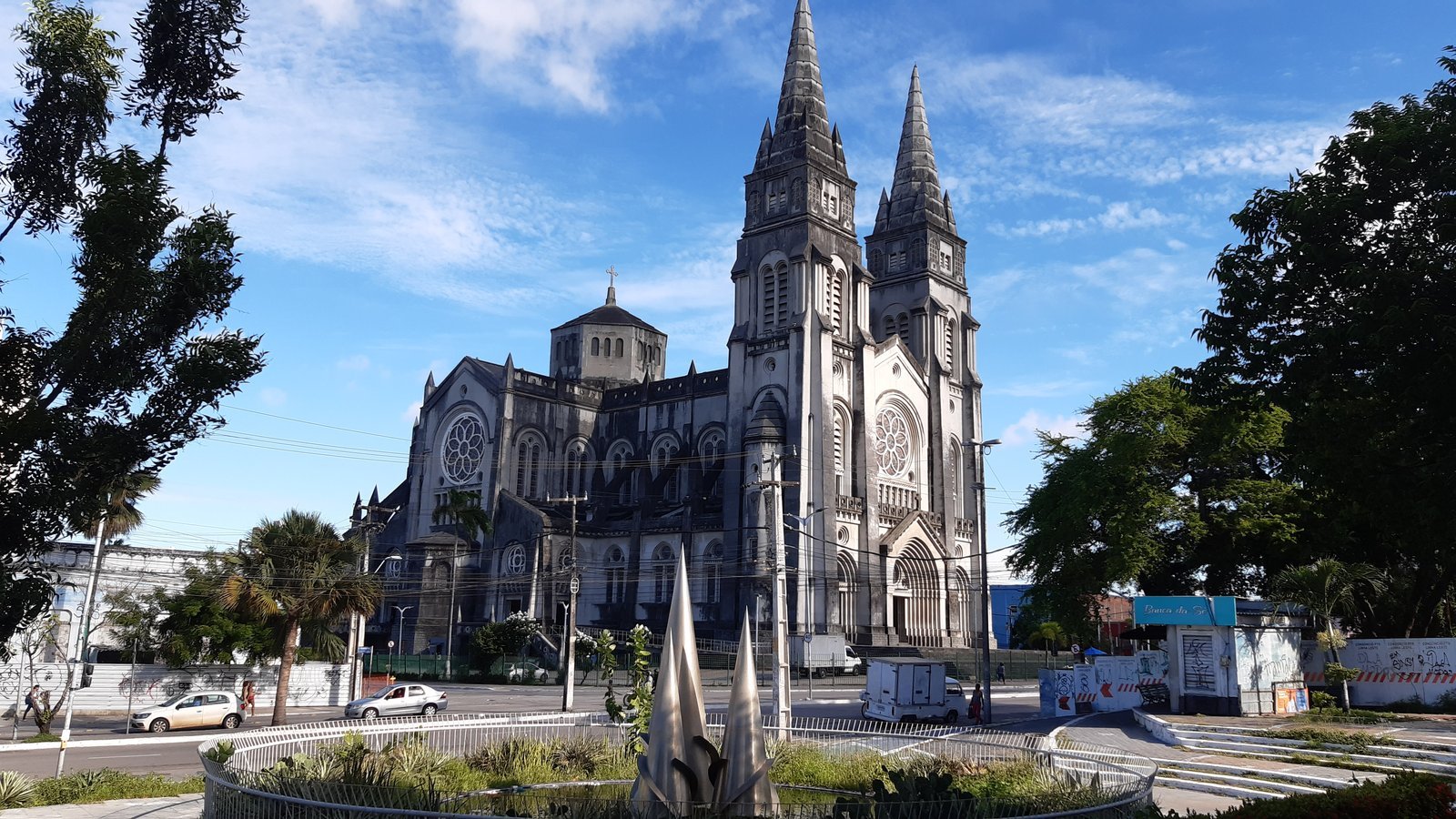 Facade of the Metropolitan Cathedral of Fortaleza, Brazil, with two pointed neo-Gothic towers seen from the tree-lined square under a blue sky