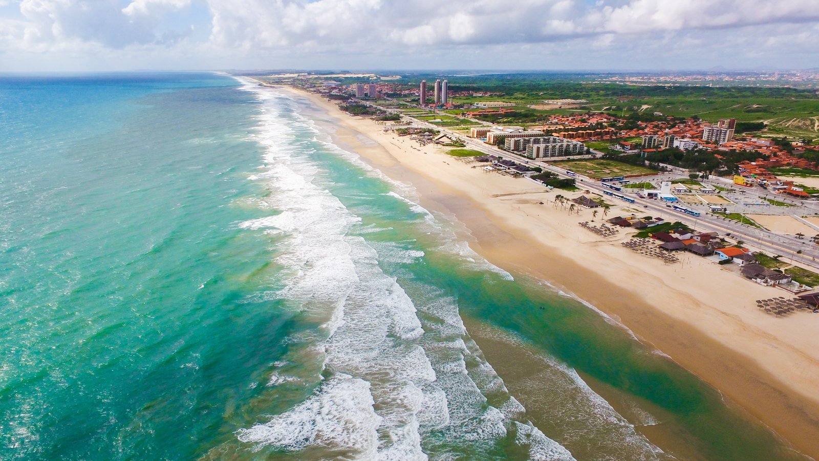 Aerial view of Praia do Futuro in Fortaleza, Brazil, with white waves breaking on a green sea, a wide strip of sand and beach clubs along the shore