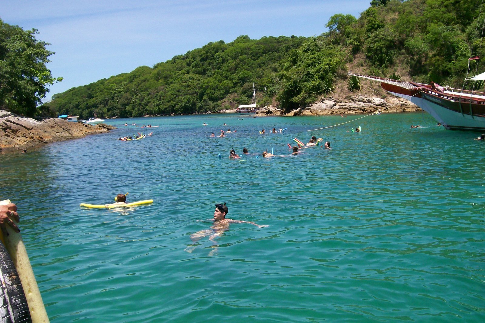 Enseada de água verde-turquesa muito transparente na Lagoa Azul, na Ilha Grande, com pessoas nadando e boiando, uma escuna ancorada à direita e costões com Mata Atlântica ao fundo