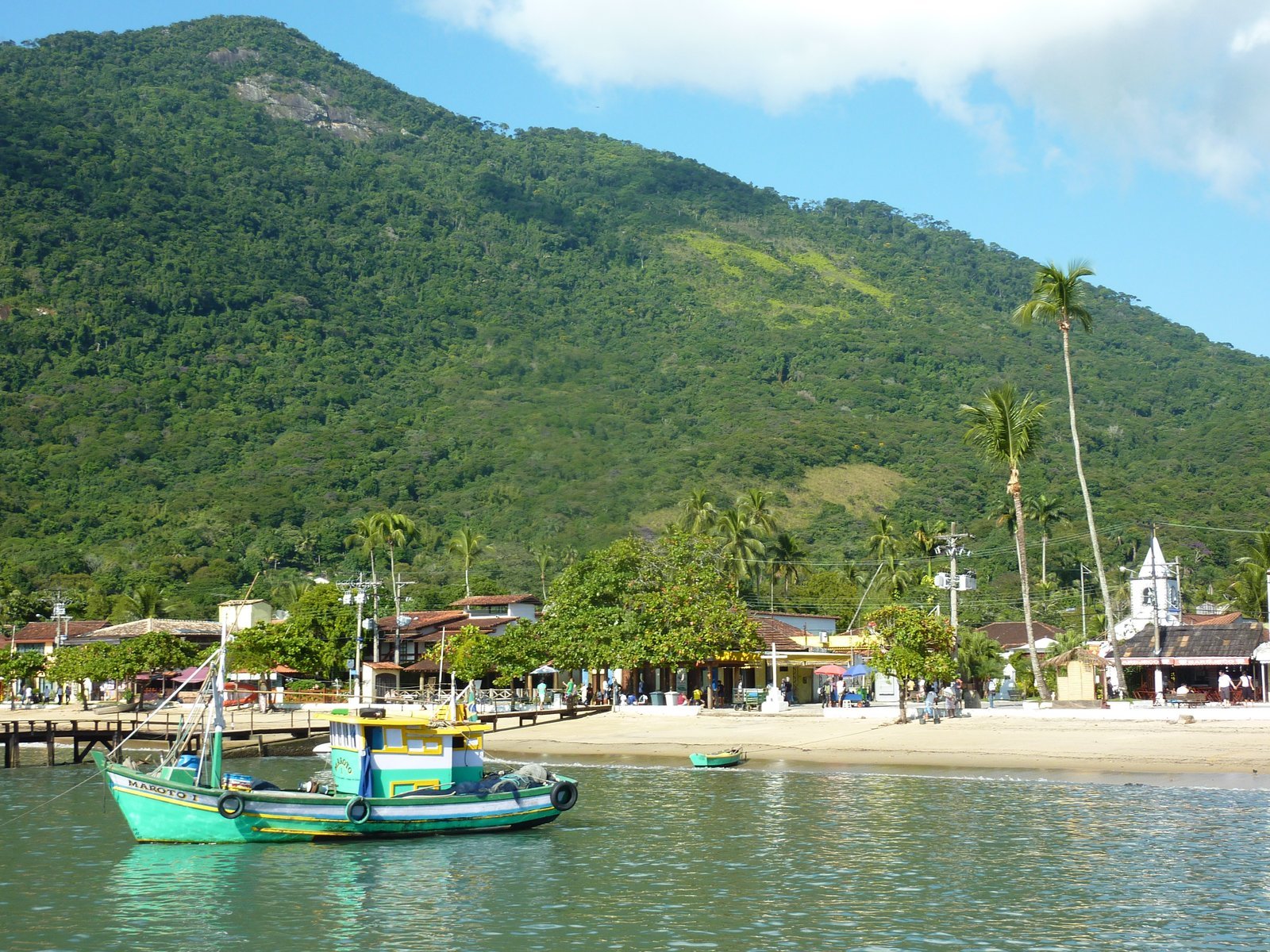 Vista da Vila do Abraão a partir do mar, com barcos coloridos ancorados na água verde, a praia, casas baixas e a igreja branca entre coqueiros, e o morro coberto de Mata Atlântica atrás