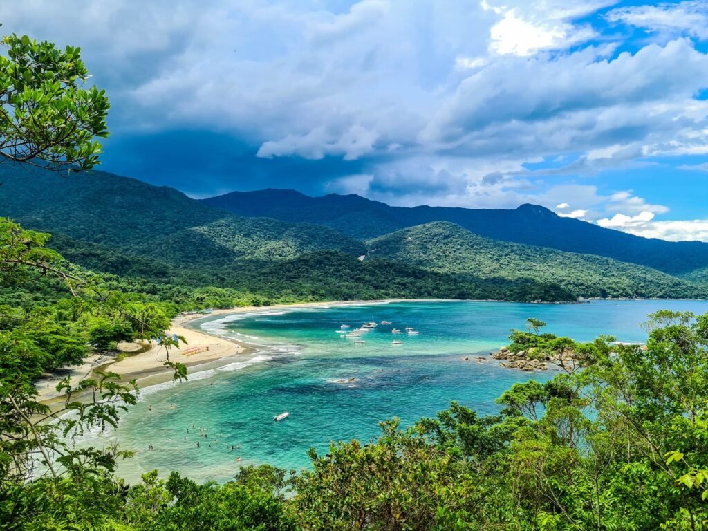 Baía de Castelhanos vista do alto, entre árvores da mata atlântica, com água verde-azulada clara, uma faixa de areia em curva e montanhas cobertas de floresta ao fundo sob céu azul com nuvens