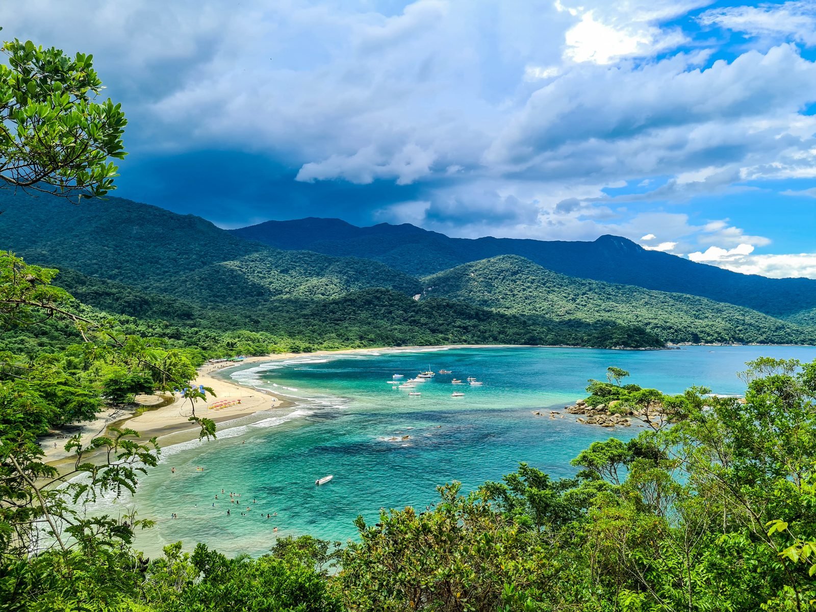Baía de Castelhanos vista do alto, entre árvores da mata atlântica, com água verde-azulada clara, uma faixa de areia em curva e montanhas cobertas de floresta ao fundo sob céu azul com nuvens