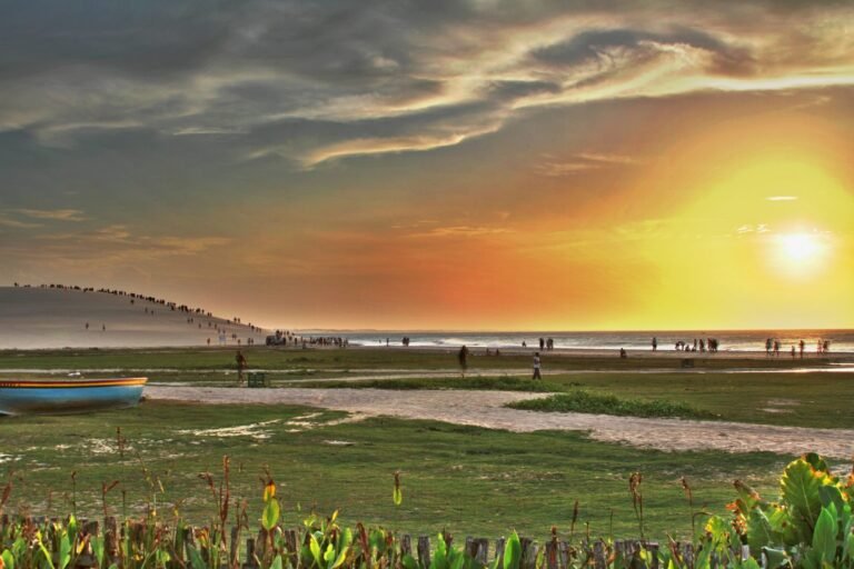 Pôr do sol alaranjado sobre a praia de Jericoacoara, com a Duna do Pôr do Sol à esquerda, pessoas caminhando na areia e um barco de pesca em primeiro plano
