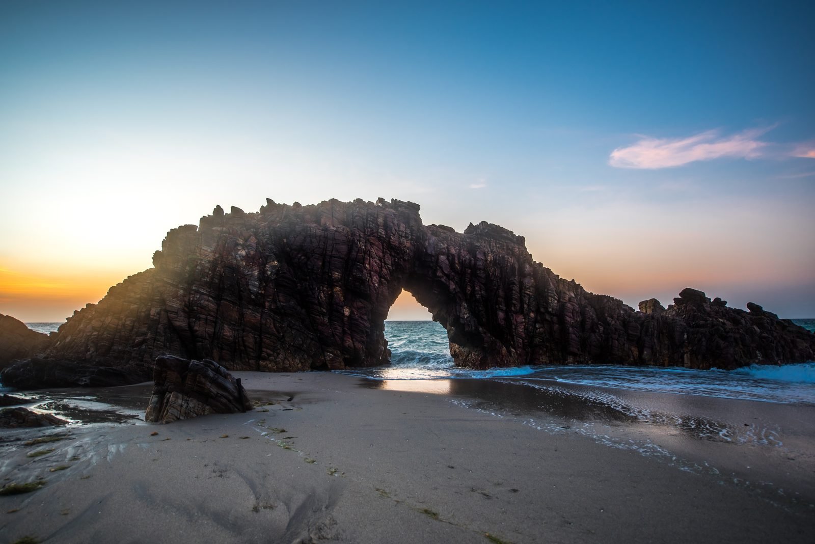 A Pedra Furada de Jericoacoara, um arco de rocha escura sobre a areia, com o céu dourado do fim de tarde visto através do buraco