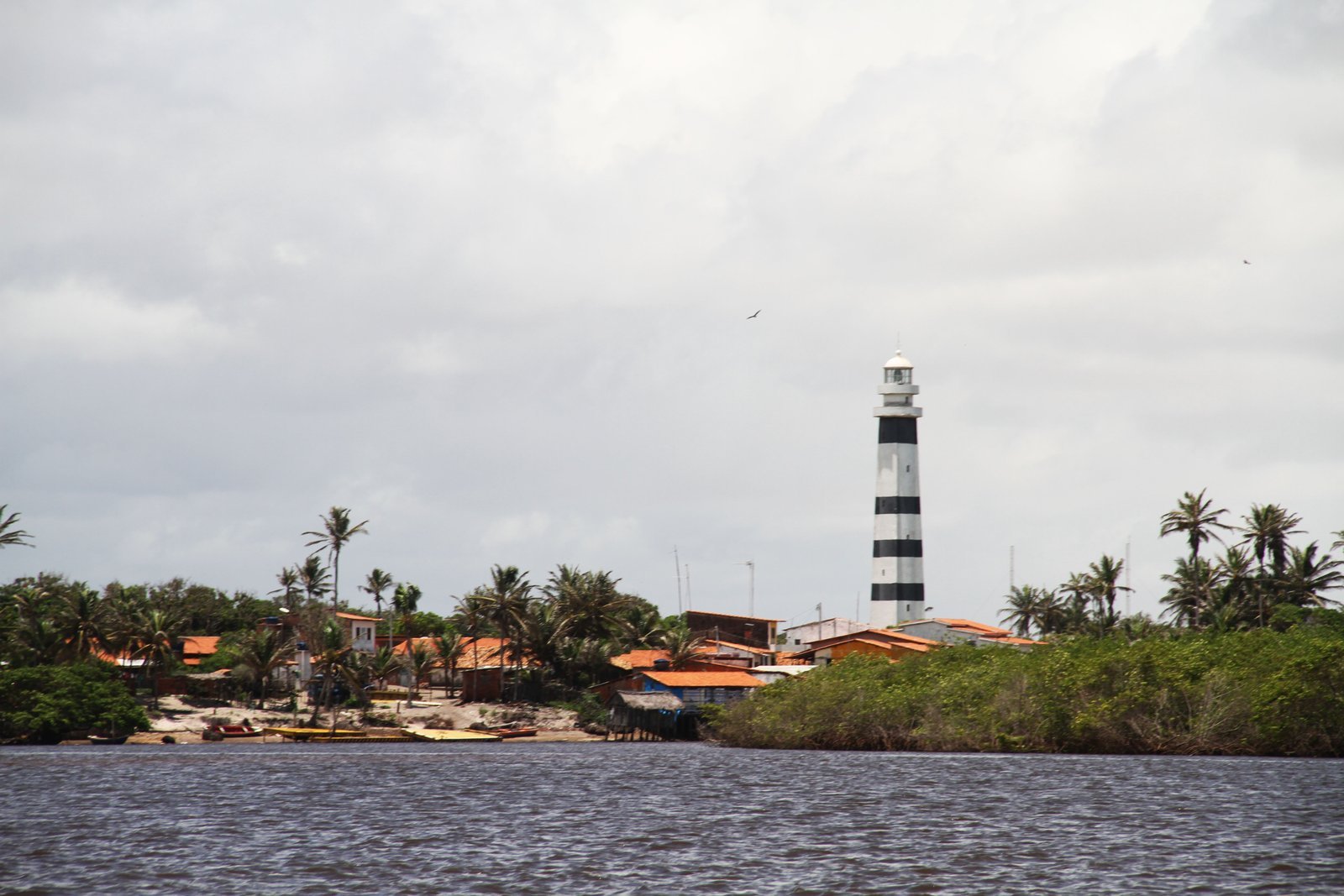 Farol Preguiças, torre listrada de preto e branco, acima das casas de telhado laranja e dos coqueiros do povoado de Mandacaru, visto do Rio Preguiças