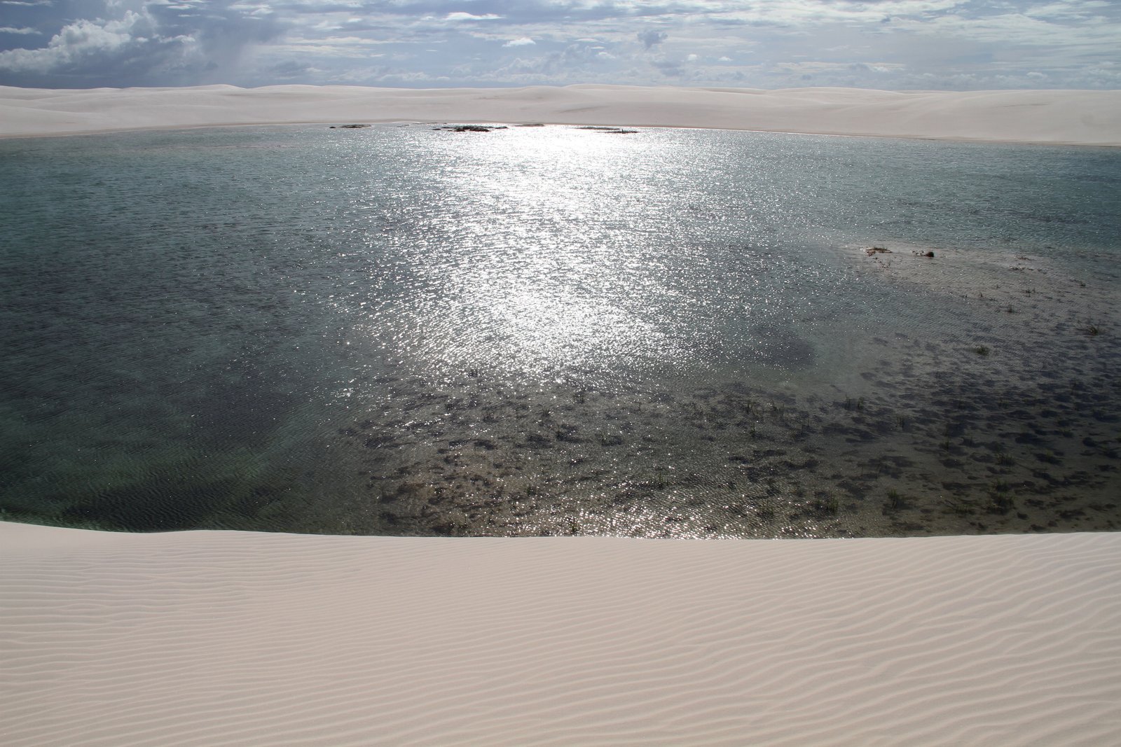 Sol refletindo na água de uma grande lagoa da Lagoa Bonita, em Barreirinhas, vista do alto de uma duna branca com marcas de vento em primeiro plano