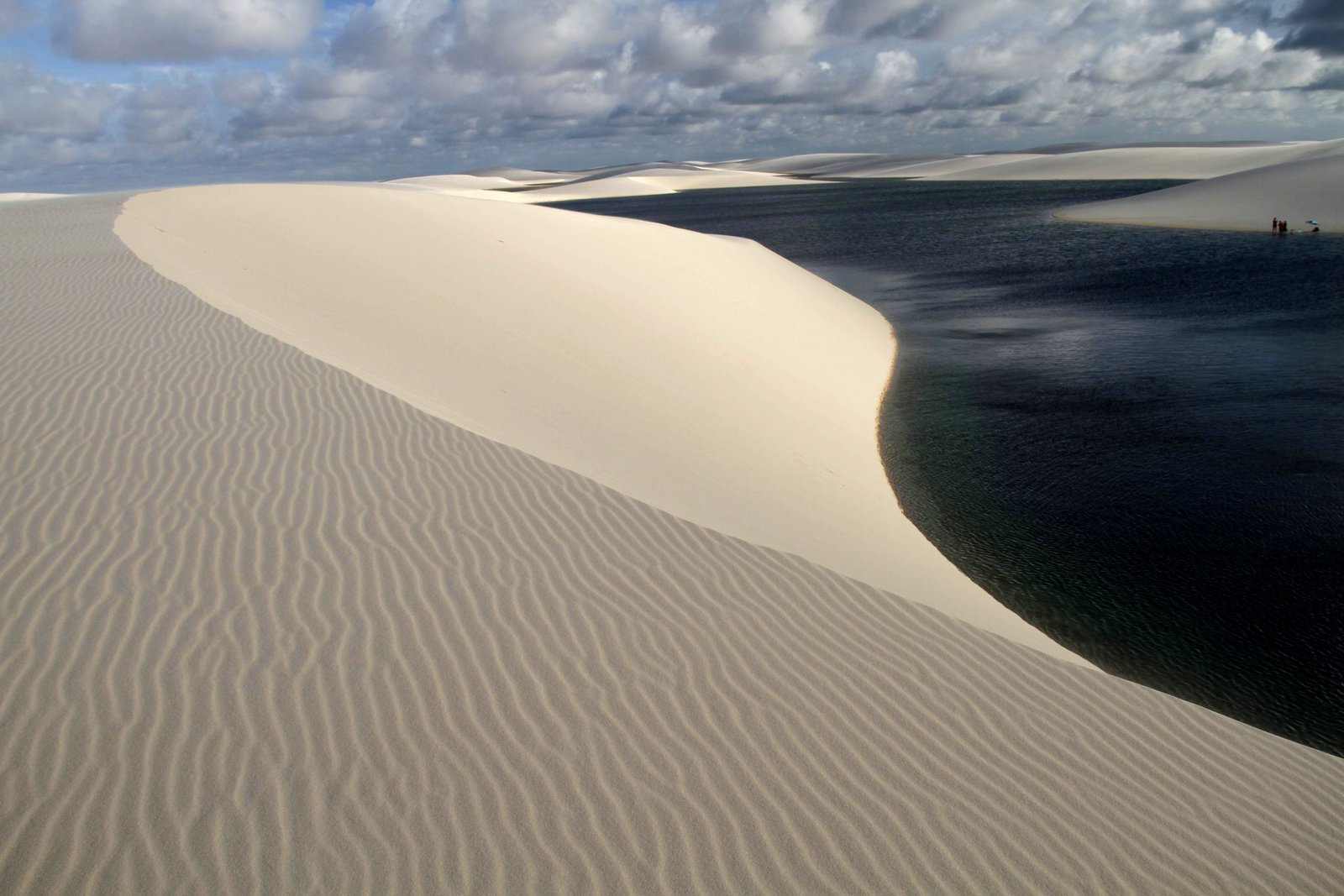 Duna branca com ondulações de vento descendo até a água escura da Lagoa das Andorinhas, em Santo Amaro do Maranhão, com pessoas pequenas na margem ao fundo