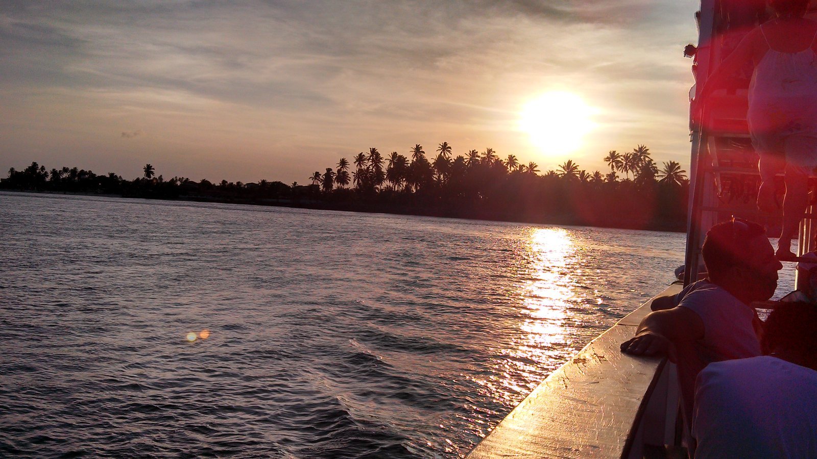 Sunset over the Mundaú lagoon in Maceió, Brazil, with palm trees on the horizon and passengers on the deck of the nine islands tour boat