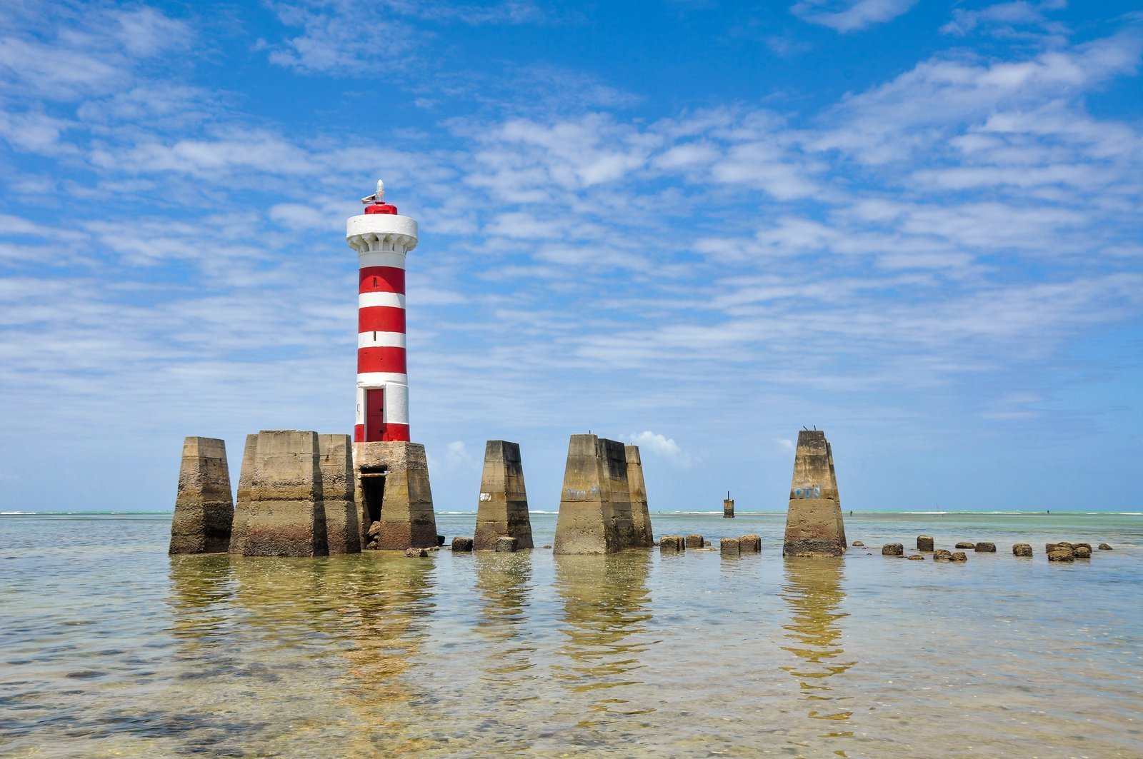 Red-and-white striped lighthouse on concrete pillars on the reef at Ponta Verde beach in Maceió, Brazil, with shallow clear water at low tide