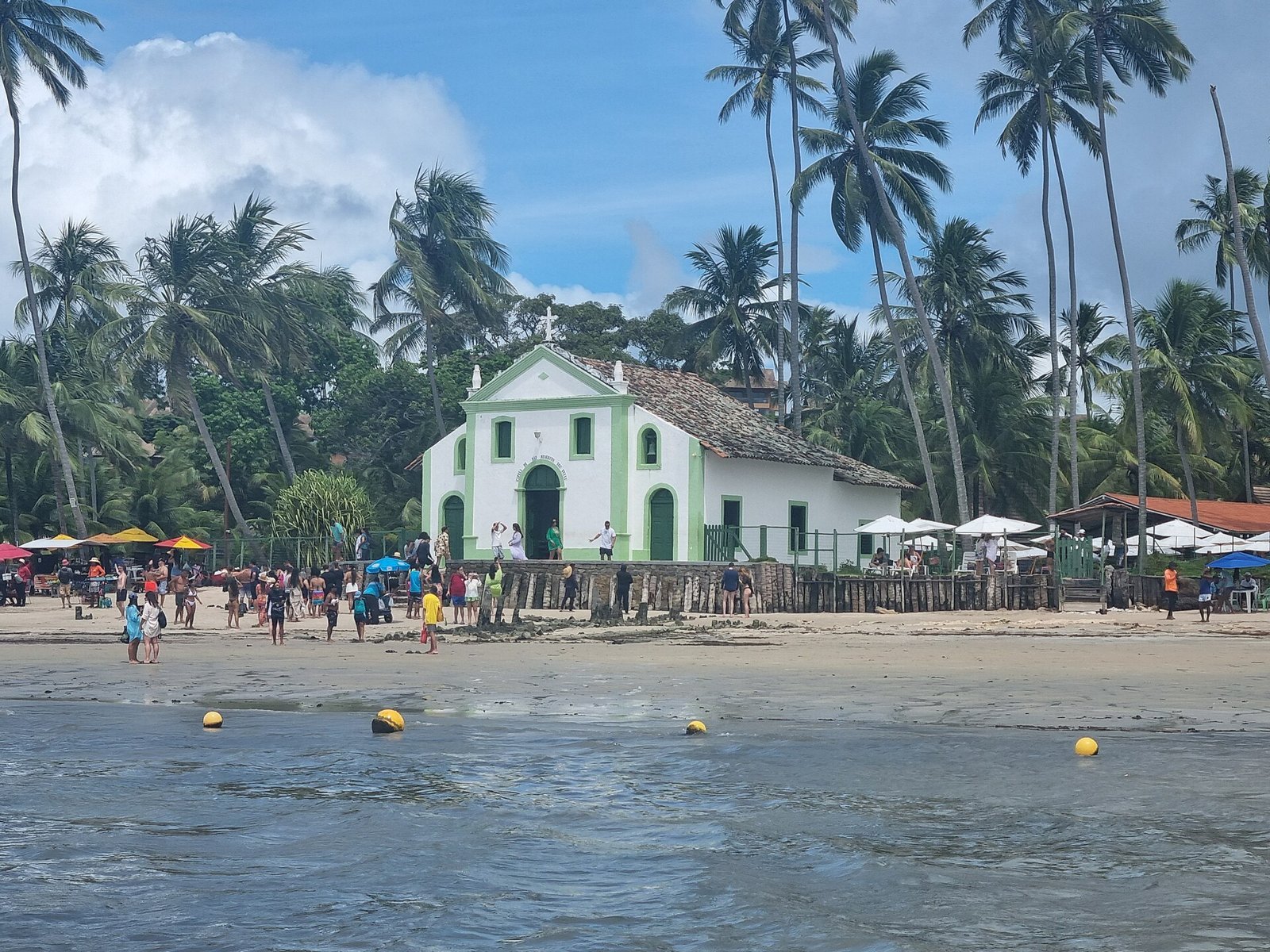 O que fazer em Maragogi: guia completo 2026 com piscinas naturais, marés, taxas, roteiro e onde ficar 5 Capela de São Benedito, pequena igreja branca com porta e janelas azuis na areia da Praia dos Carneiros, cercada de coqueiros, com banhistas ao redor e céu azul