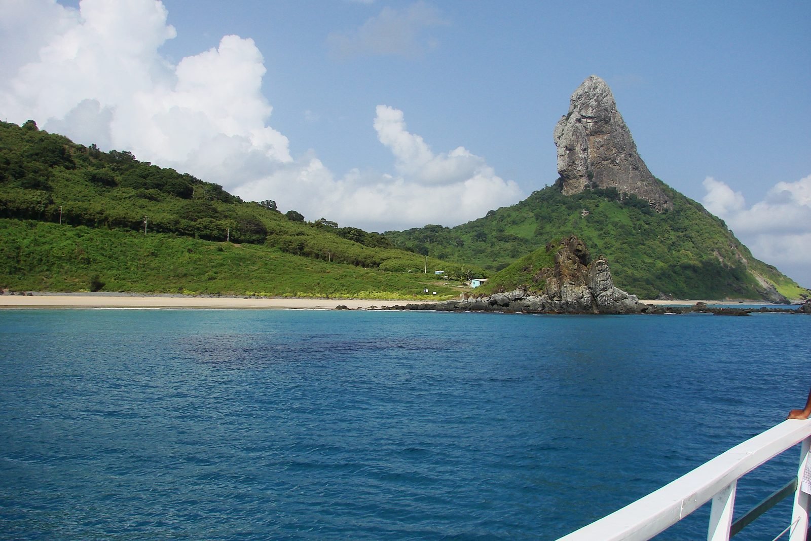 Morro do Pico, o pico rochoso mais alto de Fernando de Noronha, visto do mar, com a encosta verde e a Praia da Conceição em primeiro plano