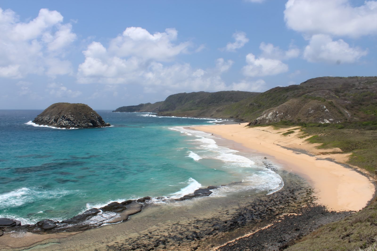 Praia do Leão em Fernando de Noronha, longa faixa de areia clara cercada de vegetação, mar azul com ondas e rochedos ao fundo, vista do alto