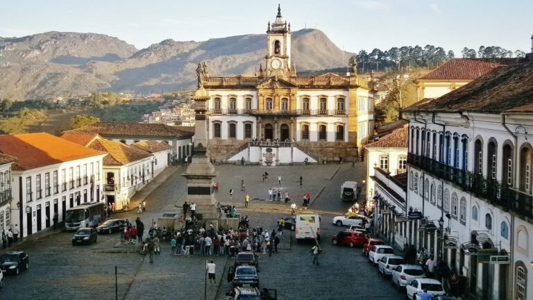 Praça Tiradentes em Ouro Preto vista do alto, com o Museu da Inconfidência e sua torre de relógio ao fundo, o monumento a Tiradentes no centro, casarões coloniais brancos dos dois lados e as montanhas ao fundo no fim da tarde