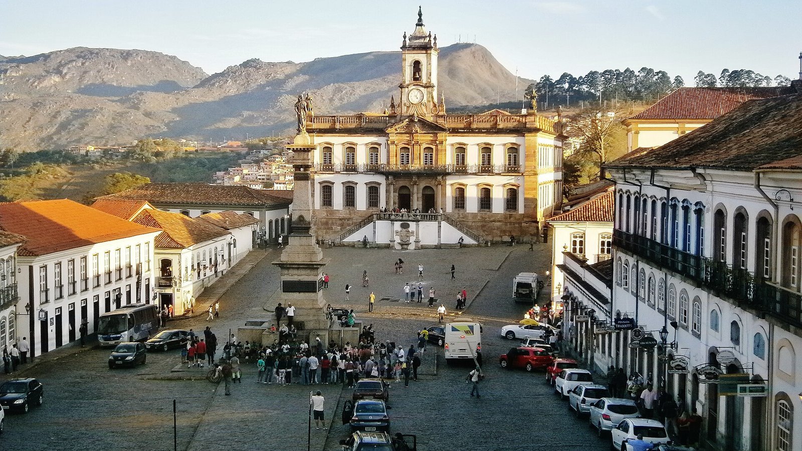 O que fazer em Ouro Preto: guia completo 2026 com história, igrejas, minas, preços, roteiro e onde ficar 2 Praça Tiradentes em Ouro Preto vista do alto, com o Museu da Inconfidência e sua torre de relógio ao fundo, o monumento a Tiradentes no centro, casarões coloniais brancos dos dois lados e as montanhas ao fundo no fim da tarde