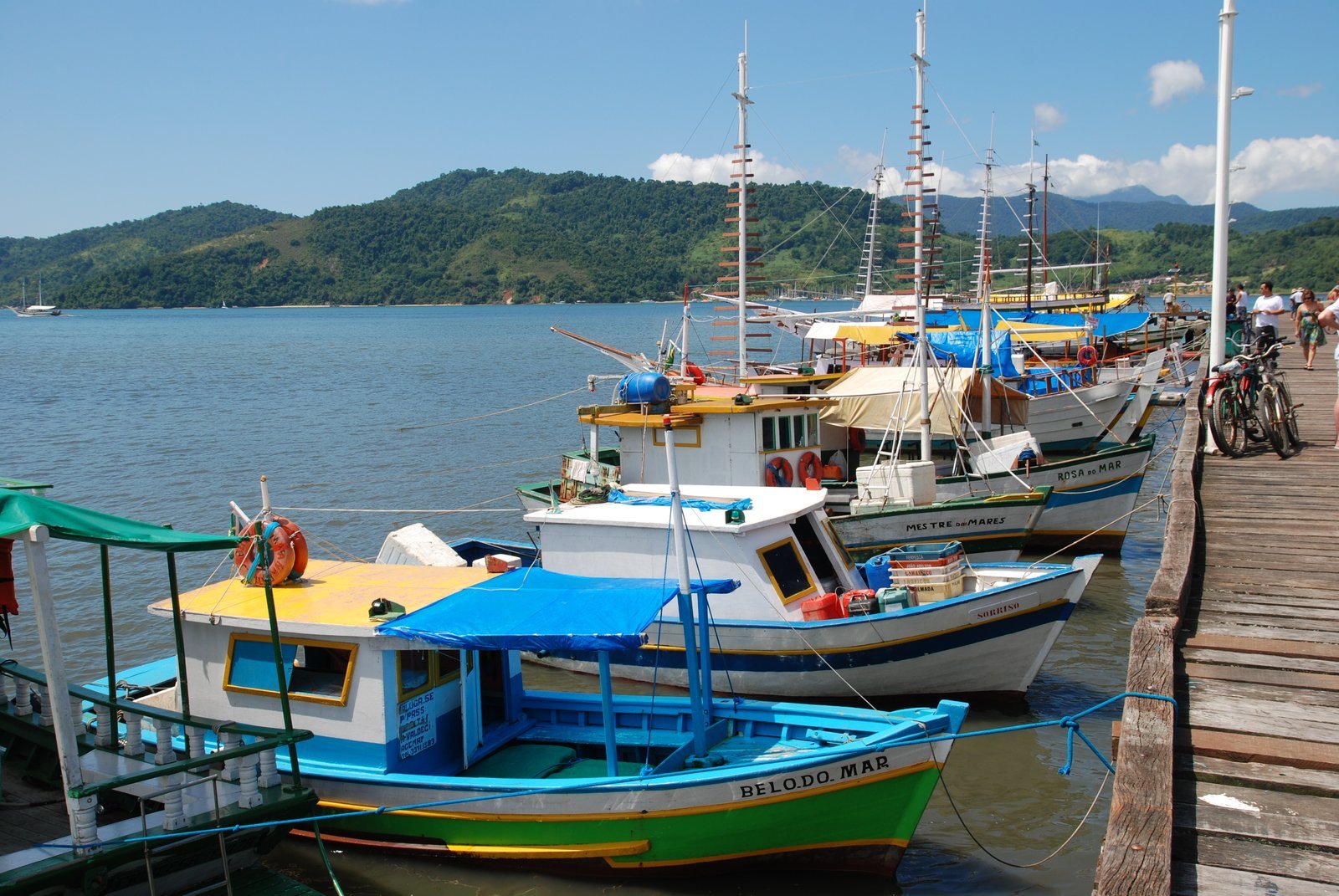 Barcos de passeio coloridos, com toldos azuis, atracados ao lado de um píer de madeira no cais de Paraty, com morros cobertos de Mata Atlântica do outro lado da baía