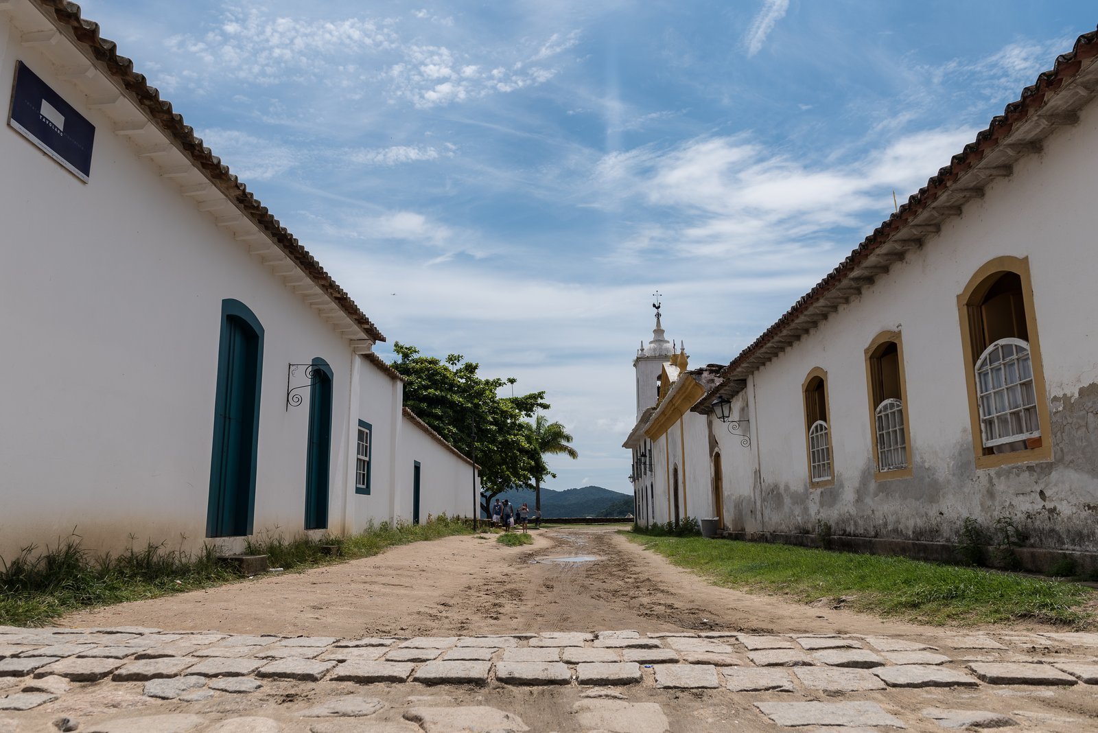 Rua de pedras irregulares no centro histórico de Paraty, ladeada por casas coloniais brancas de portas e janelas coloridas, com uma torre de igreja ao fundo e céu azul