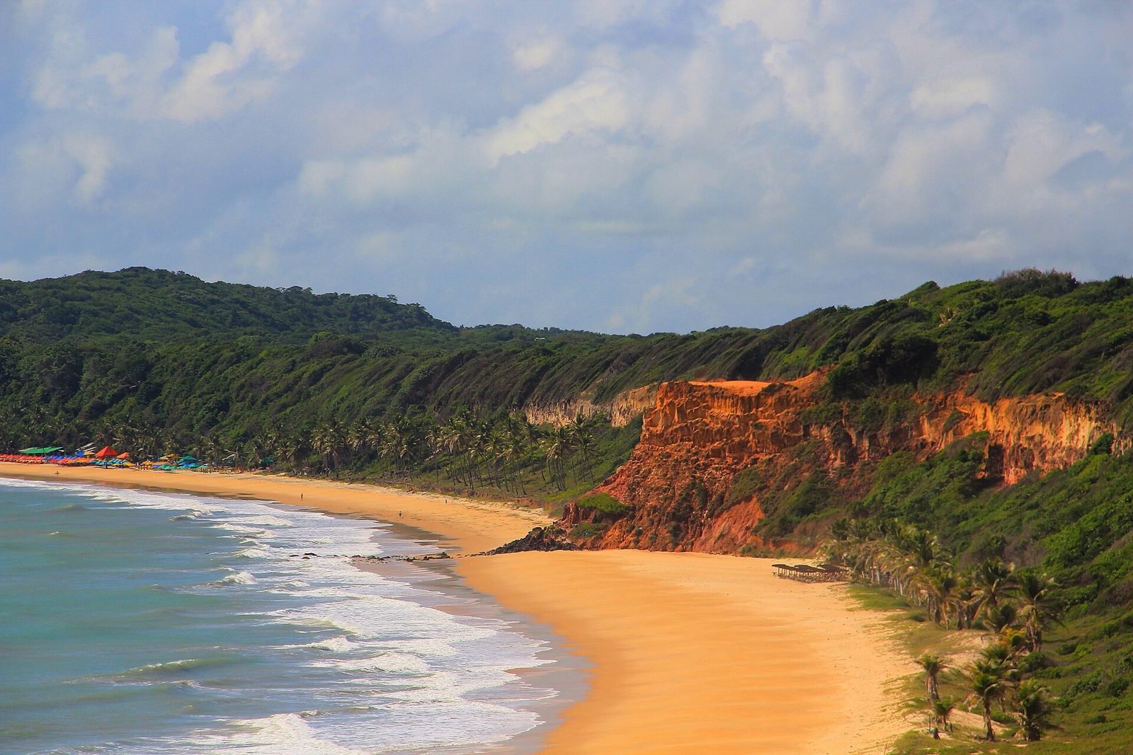 Vista do alto de uma longa praia de areia dourada, com mar de ondas brancas à esquerda, uma falésia alaranjada no centro e morros cobertos de mata verde e coqueiros, com guarda-sóis coloridos ao fundo