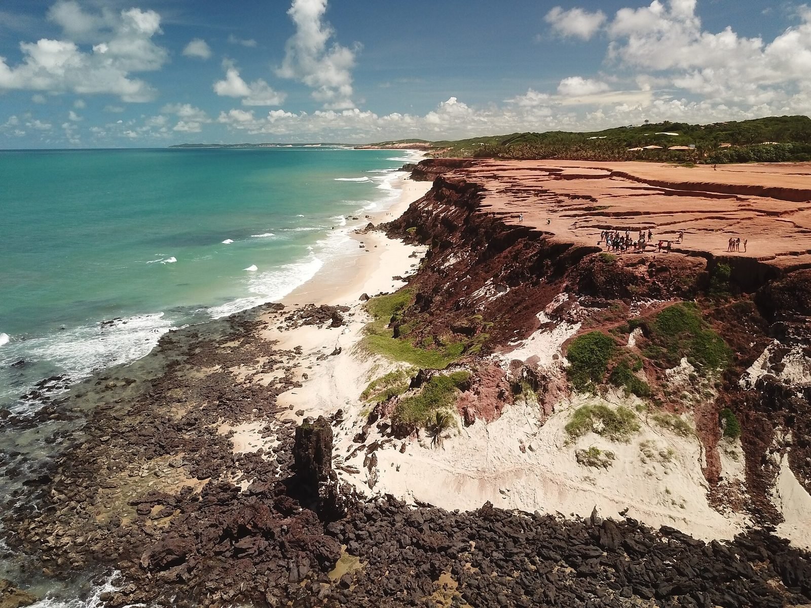 Vista aérea do Chapadão, um platô de falésias vermelhas com pessoas na borda, descendo até pedras escuras e uma faixa de areia clara, com o mar verde-azulado à esquerda e a mata ao fundo