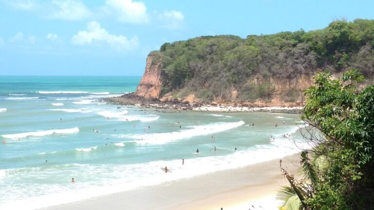 Praia em Pipa vista do alto, com mar azul-turquesa, ondas pequenas, surfistas e banhistas na água, faixa de areia clara à direita e uma falésia alaranjada coberta de mata verde avançando sobre o mar
