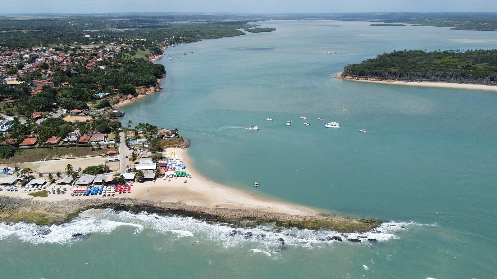 Vista aérea da ponta de areia de Tibau do Sul, com barracas e guarda-sóis coloridos, a cidade com telhados vermelhos à esquerda, barcos ancorados na água esverdeada da Lagoa de Guaraíras e uma ilha de mata à direita