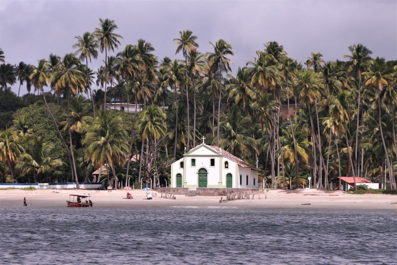 Pequena igreja branca com detalhes azuis de São Benedito na areia da Praia dos Carneiros, cercada de coqueiros, com o mar verde e calmo à frente