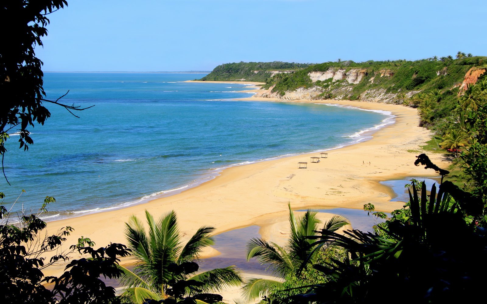 Praia do Espelho, no litoral de Porto Seguro (BA), vista do alto da falésia, com mar azul-turquesa, faixa de areia dourada curva e vegetação verde em primeiro plano