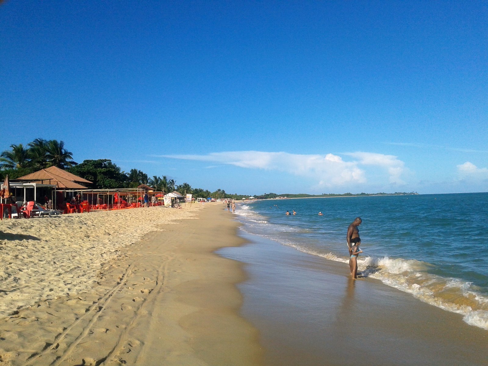 Faixa larga de areia clara da Praia de Taperapuã, em Porto Seguro (BA), com mar azul de ondas baixas e barracas de praia cobertas de palha ao fundo