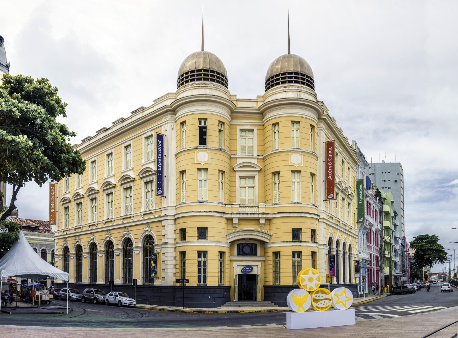Yellow two-story historic building with twin domes on Marco Zero square in Recife Antigo, with a letter sculpture on the sidewalk under a blue sky with clouds