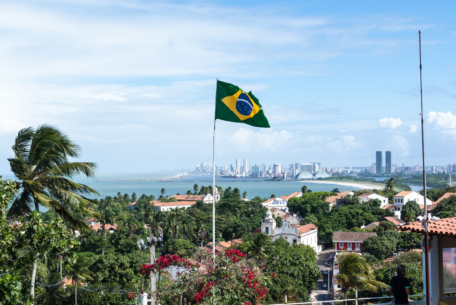 View from Alto da Sé in Olinda, with a Brazilian flag on a pole, coconut palms, houses among flowering trees and the high-rises of Recife on the horizon by the sea