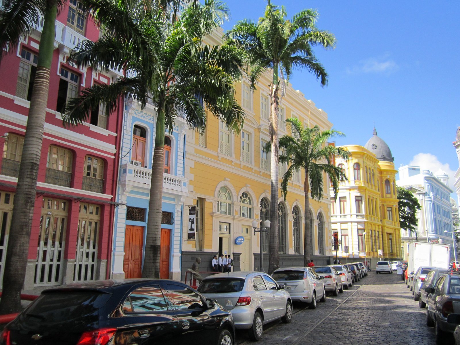 Rua do Bom Jesus in Recife Antigo, with colorful pink, blue and yellow townhouses, tall palm trees, parked cars and a dome in the background under a blue sky
