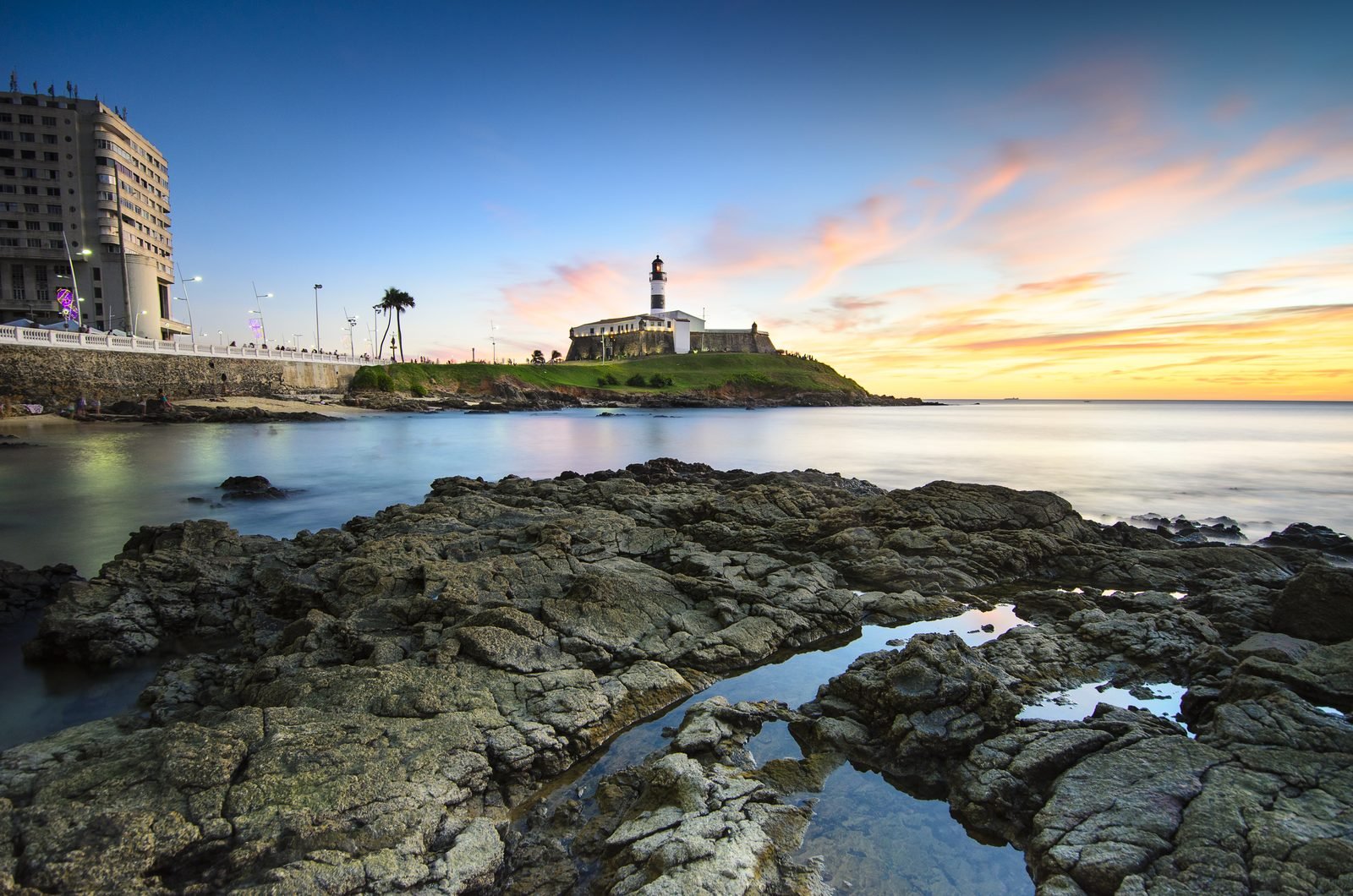 Farol da Barra lighthouse in Salvador, Brazil, at dusk, with the lit fort in the distance, an orange sky and rocks along the shore