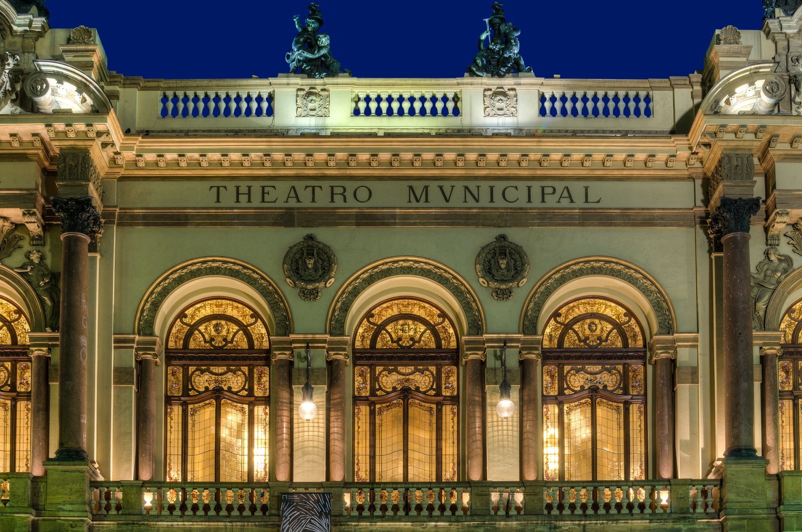 Facade of the Theatro Municipal in São Paulo lit up at dusk, with arched windows, columns and the Theatro Municipal lettering
