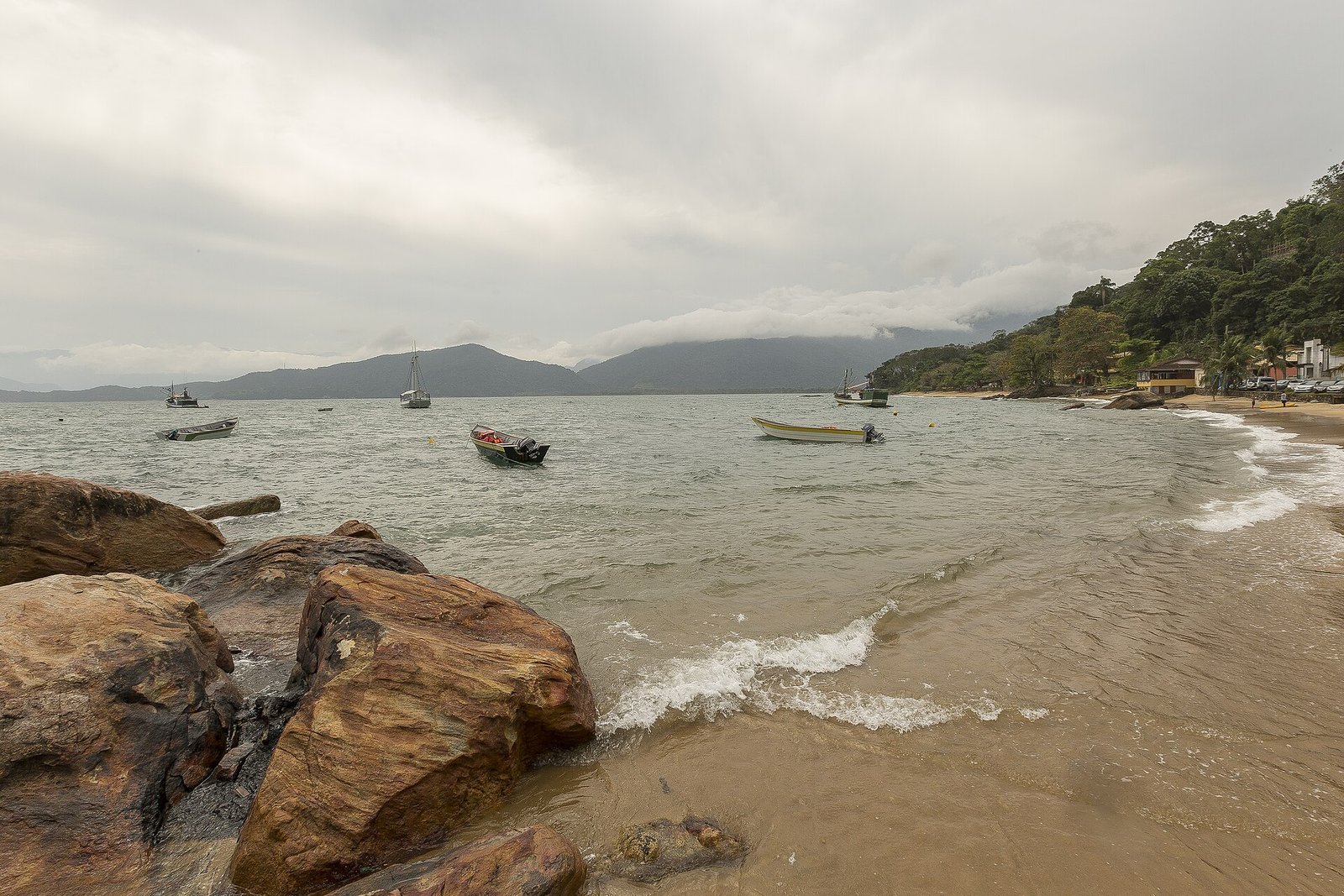 Barcos de pesca coloridos ancorados em frente à pequena praia da vila de Picinguaba, em Ubatuba, com casas e morros de mata atlântica ao fundo em dia nublado